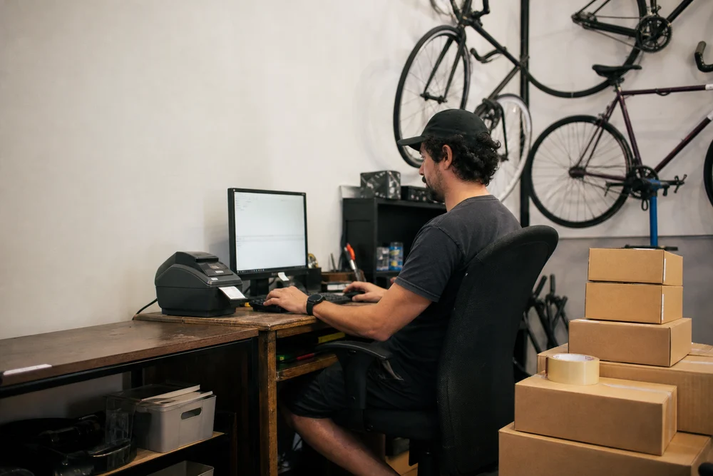 Dono de uma loja de peças de bicicleta emitindo a nota fiscal de um pedido no computador da sala dos fundos, com a impressora de etiquetas e caixas prontas ao lado